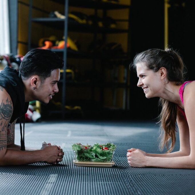 man and woman at the gym doing planks while looking at each other and smiling